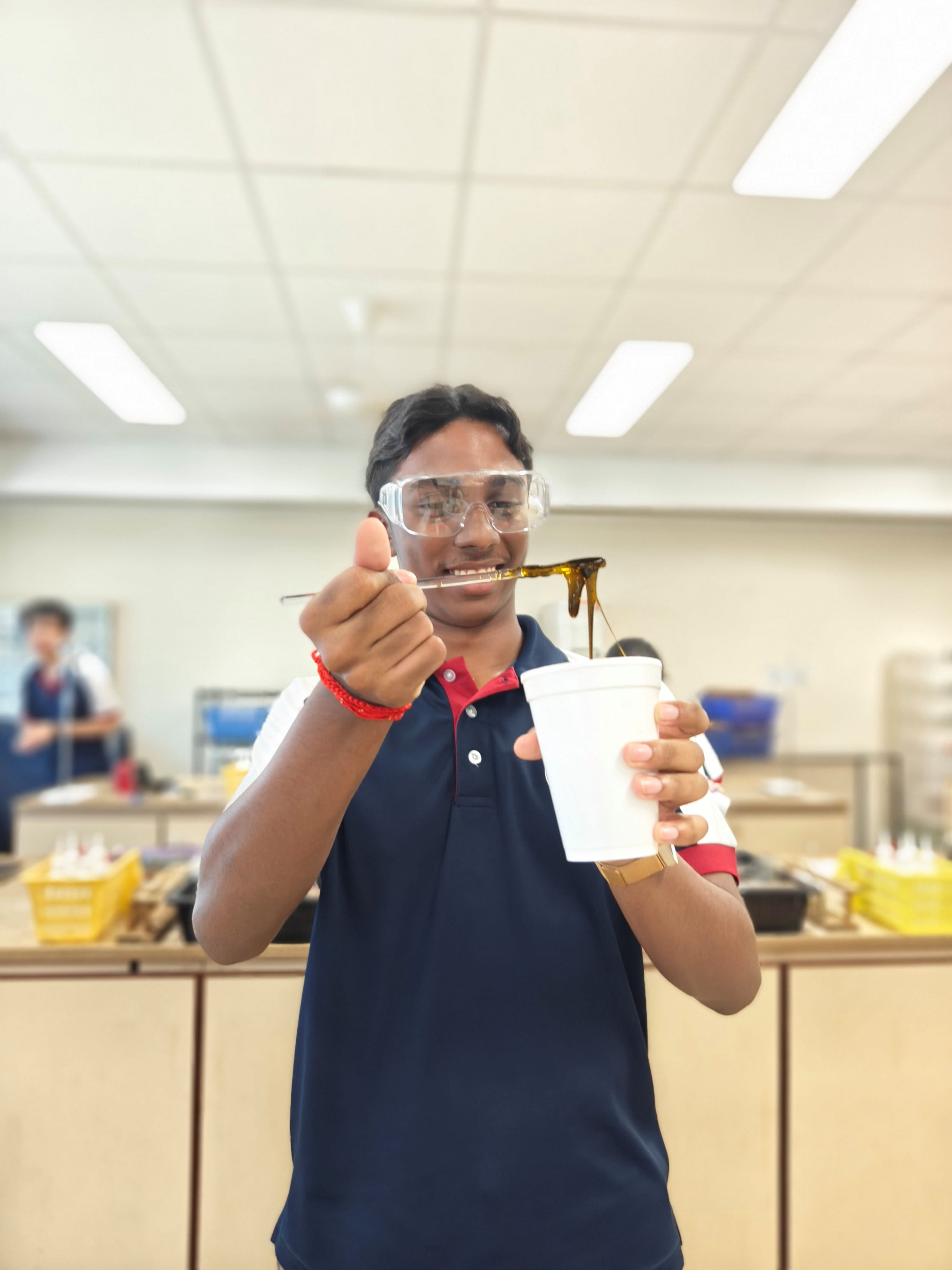 A male student in a navy blue polo shirt and safety goggles grins at the camera while holding a white cup in one hand and stretching a dark, sticky polymer substance with the other.